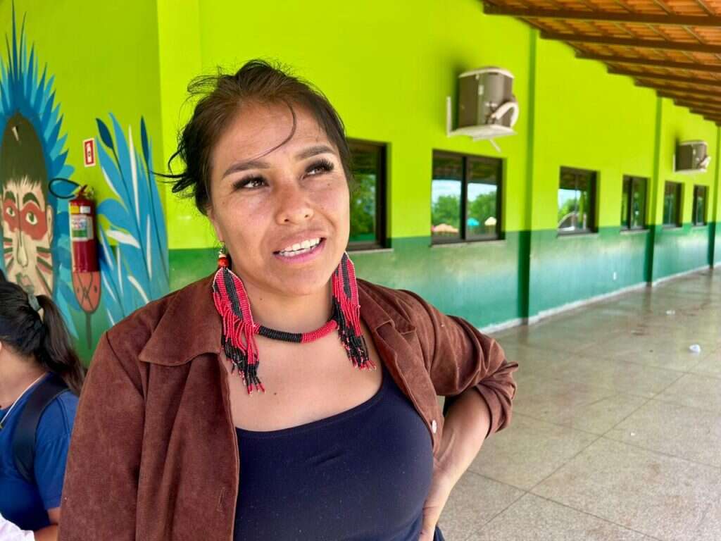 Uma mulher indígena sorrindo e olhando para cima, com brincos e colar de contas pretas e vermelhas longos. Ela veste uma blusa preta e um casaco marrom, e está em frente a uma parede verde com uma pintura mural.