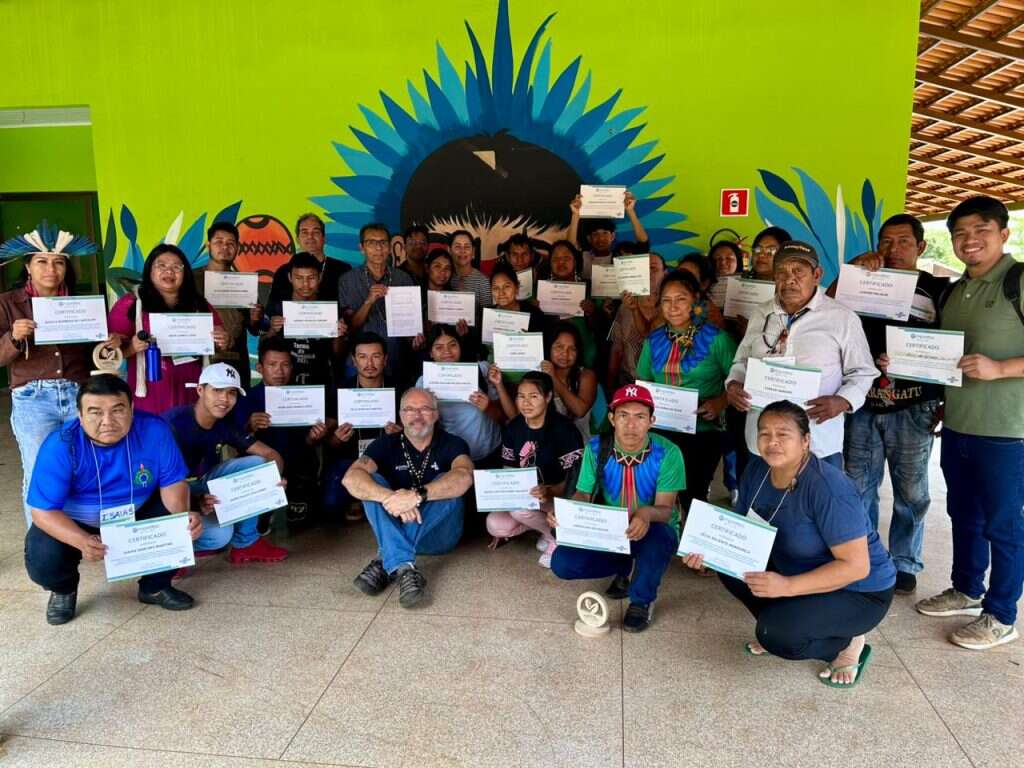 Foto de grupo de aproximadamente 30 participantes, a maioria deles indígenas, em frente a uma parede verde com grafismos artísticos em azul e preto. Todos seguram certificados do curso Empretec. Há homens e mulheres, alguns trajados com adornos tradicionais, celebrando o encerramento do programa.