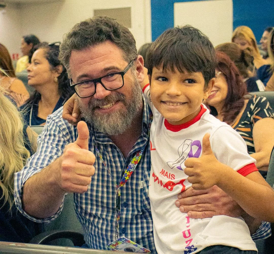 Deputado federal Marcos Pollon sorri ao lado de uma criança, ambos fazendo sinal de positivo, em evento de apoio a famílias neurodivergentes.